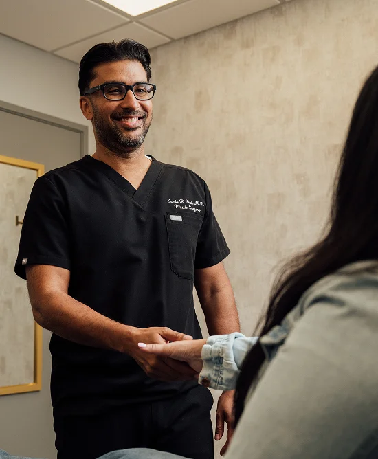 Shot of Dr. Samir Shah wearing black scrubs, and glasses, shaking a clients hand.