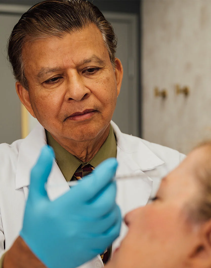Dr. Shah wearing a white lab coat and blue gloves is administering an injection to the temple area of a blonde patient. - Botox and Wrinkle Relaxers in Orlando Park, IL