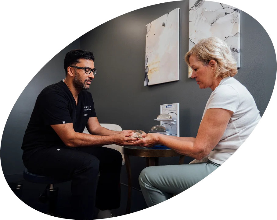Samir Shah, MD, a man wearing black scrubs and glasses, is sitting across a table from a female patient. He is holding out a breast implant sizer to her, and other sizers are visible in a display rack between them.