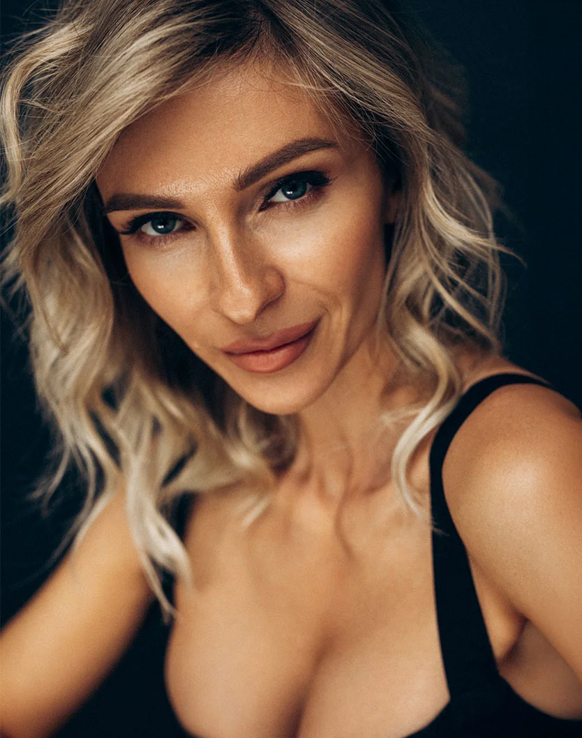 Light-skinned woman with wavy blonde hair. She wears a dark sleeveless top and gazes directly at the viewer with a neutral expression. A solid black background contrasts with her light features.- Breast Lift in Orlando Park, IL