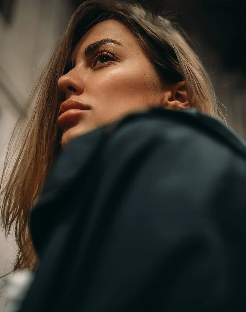 A low-angle shot of a beautiful young woman looking up and to the side, with a determined or pensive expression. She has long, light brown hair and is wearing a dark jacket. The background is blurred, suggesting an urban or modern setting, possibly indoors. The lighting highlights her face, focusing on her brow, eyes, and cheekbones. - Brow Lift in Orlando Park, IL