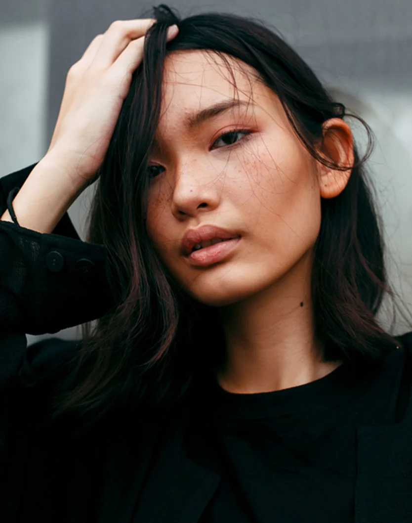 A close-up portrait of a woman with dark, long hair, wearing a black top, running her hand through her hair and looking directly at the camera. - Ear Reshaping in Orlando Park, IL