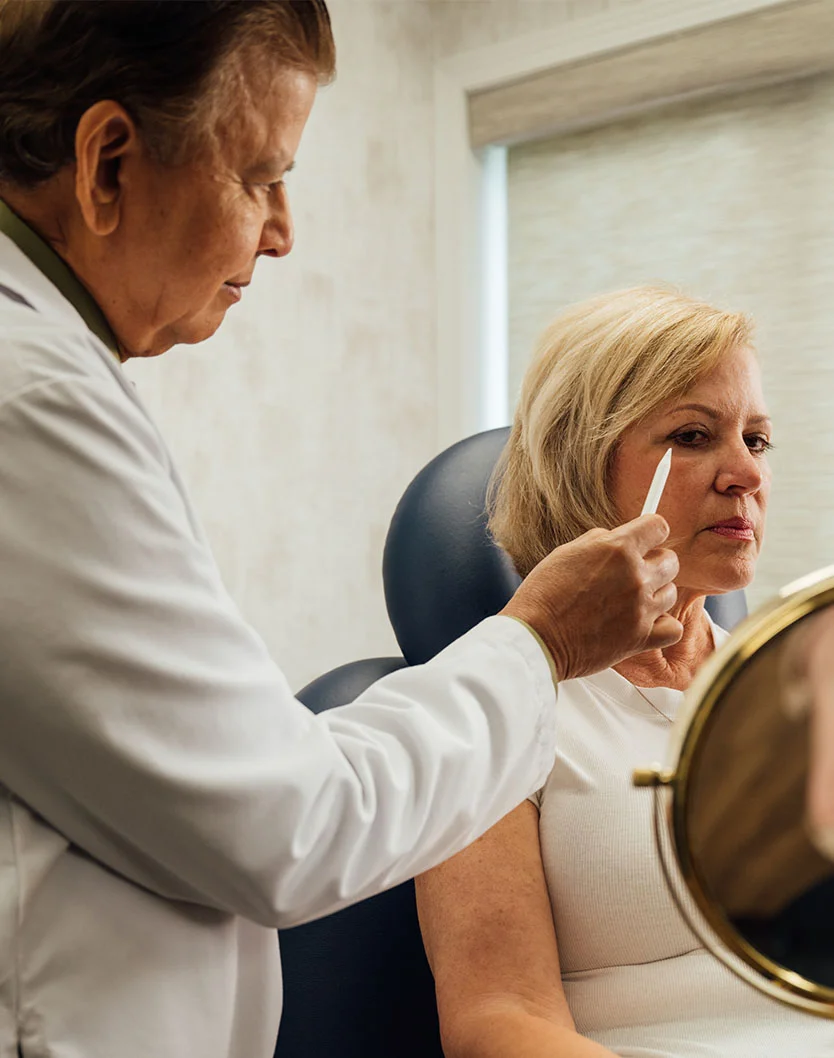 A plastic surgeon, Dr. Shah, in a white lab coat, examines a blonde female patient's eye with a small white tool while she looks in a handheld mirror. - Eyelid Surgery in Orlando Park, IL