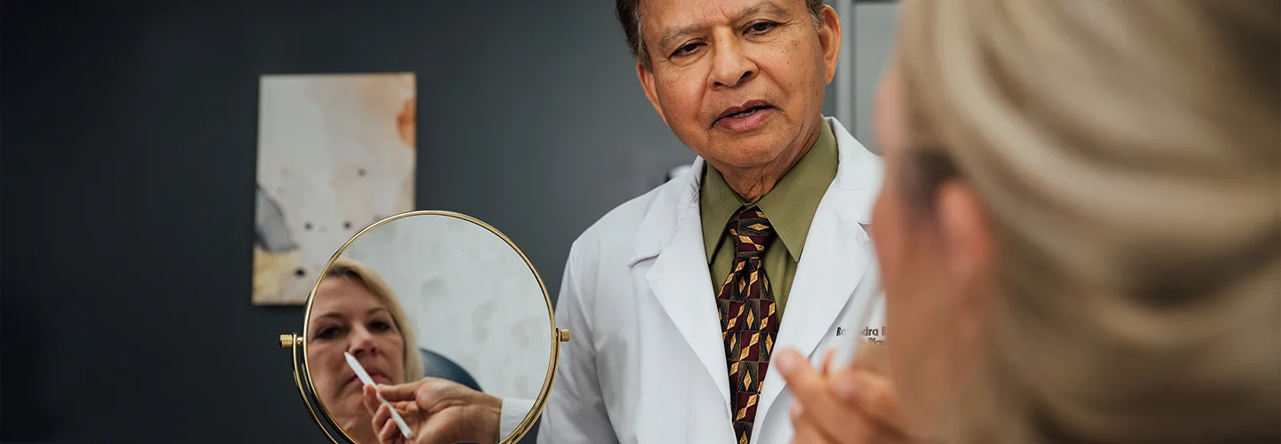 Rajendra Shah, MD, a man wearing a white lab coat, green shirt, and patterned tie, is looking down at a female patient holding a hand mirror. The patient's face is reflected in the mirror, where another hand is gently touching her face.