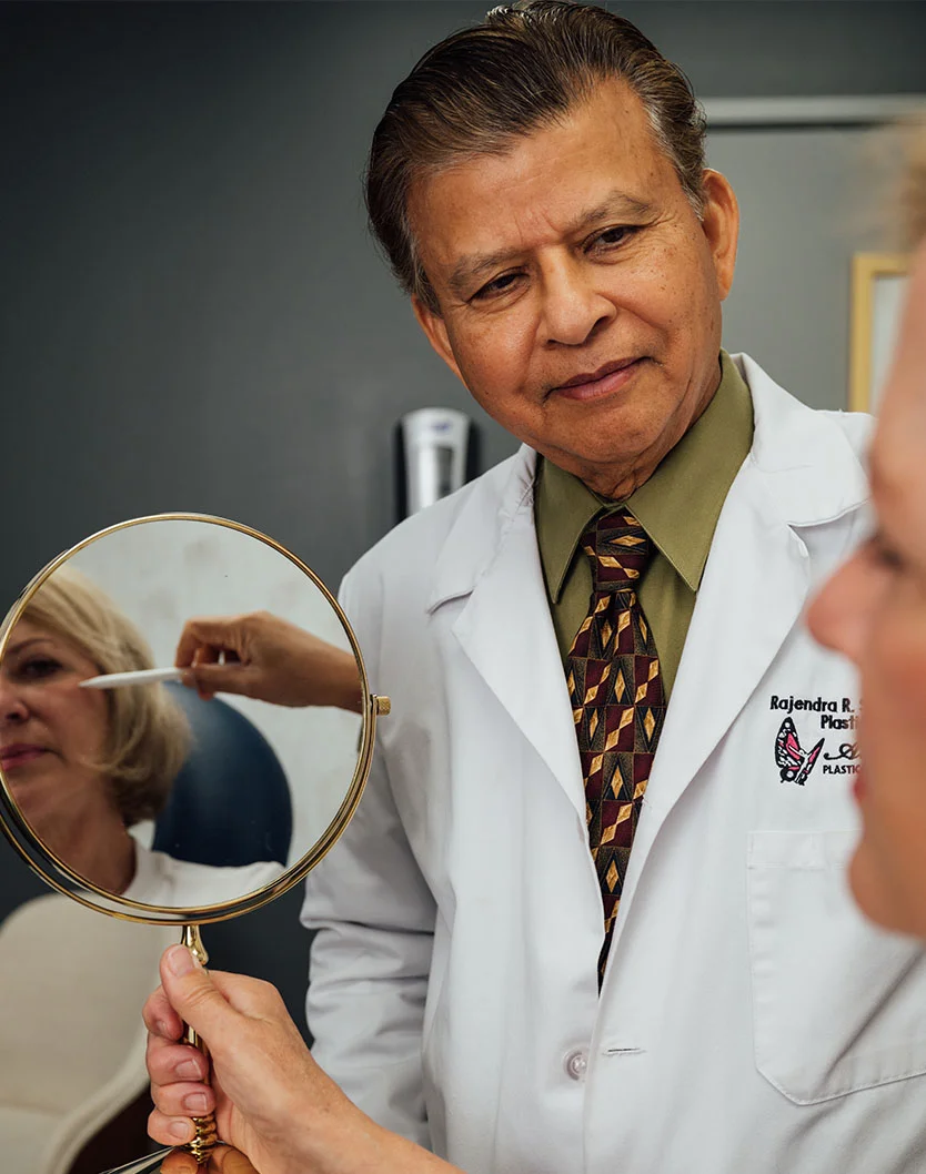 Rajendra Shah, MD, a man wearing a white lab coat, green shirt, and patterned tie, is looking down at a female patient holding a hand mirror. The patient's face is reflected in the mirror, where another hand is gently touching her face. - Facial Fat Grafting in Orlando Park, IL