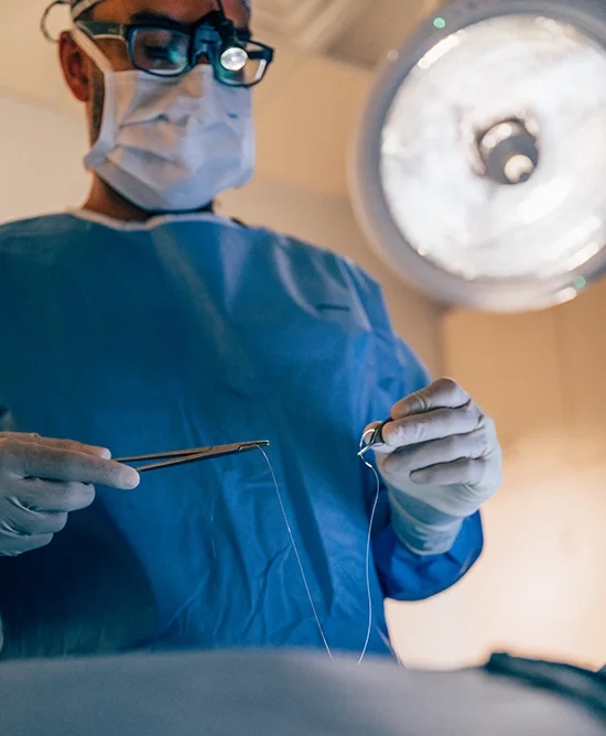A close-up of Samir Shah, MD, wearing a surgical mask, glasses, and a blue surgical gown, holding a needle and thread with surgical tools while performing a procedure under a bright overhead light.