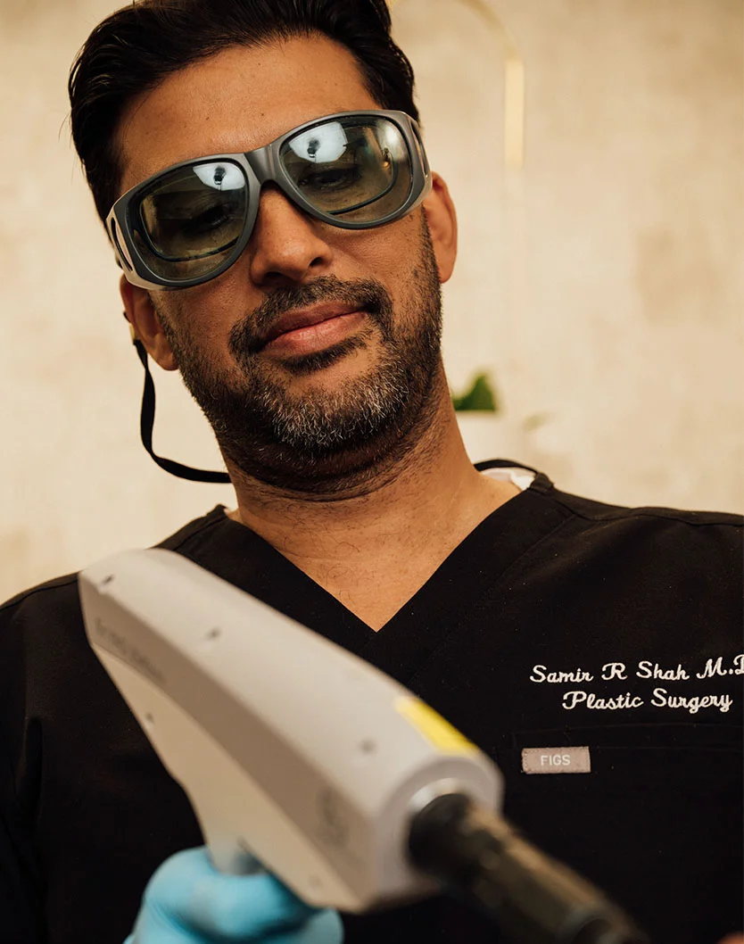A close-up of Dr. Samir Shah, MD, a man with a beard, wearing protective glasses and a black scrub top embroidered with "Samir R Shah M.D. Plastic Surgery." He is holding a laser device and treating a patient who is lying down. - Laser Skin Resurfacing in Orlando Park, IL