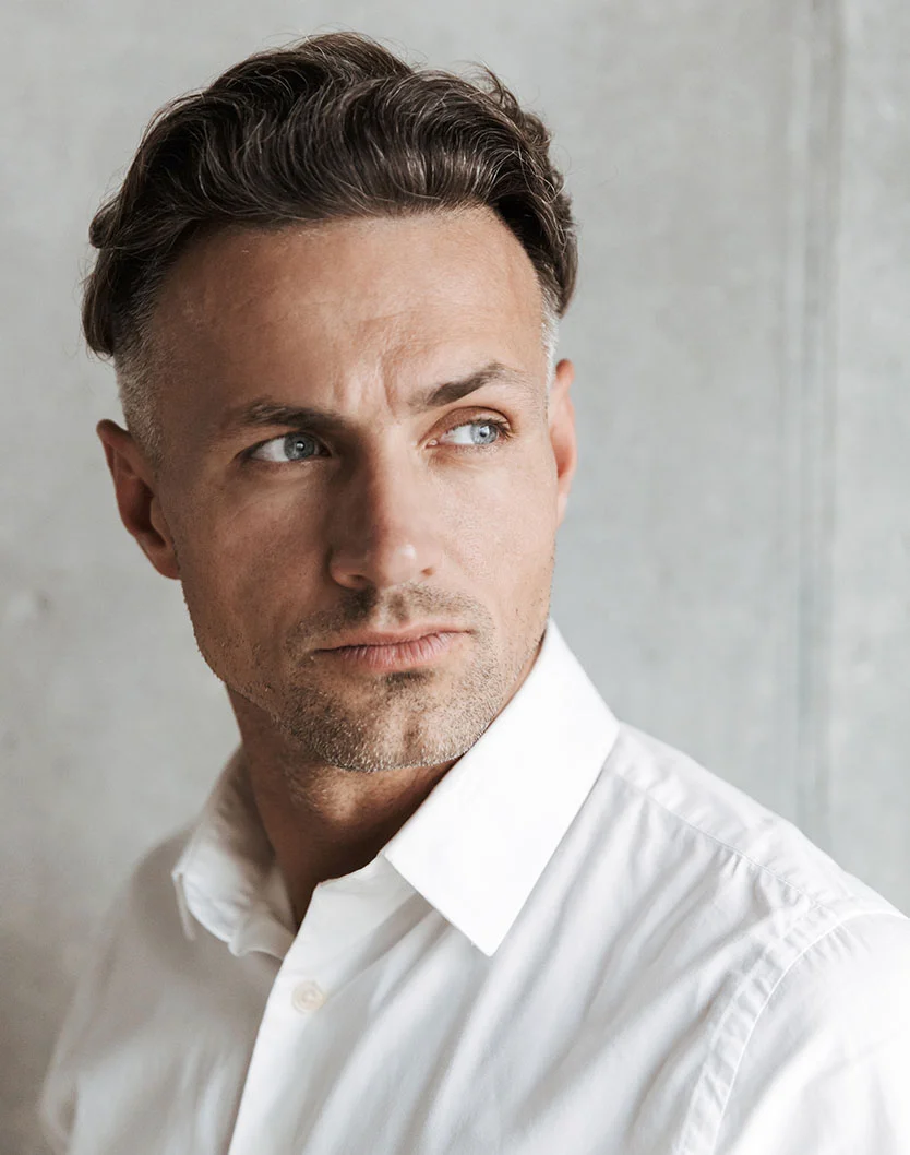 A man with dark and graying hair, wearing a white collared shirt, looking thoughtfully to his left. He has a serious expression on his face and is standing against a gray wall. - Male Facelift in Orlando Park, IL
