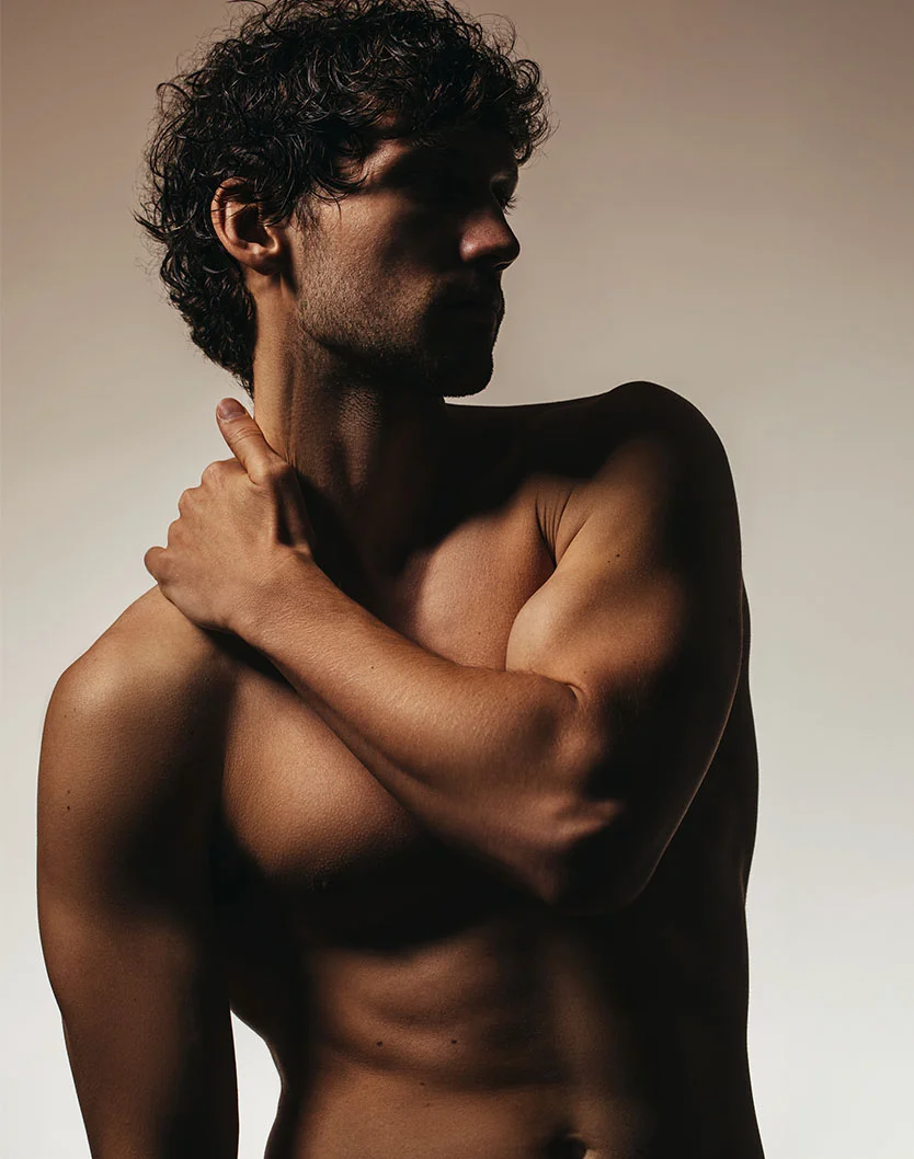 A moody, high-contrast portrait of a shirtless, muscular man with dark, curly hair. He is positioned in profile against a light background, but his face and body are mostly in shadow, highlighting the definition in his arm as he rests his hand on his opposite shoulder. - Male Liposuction in Orlando Park, IL