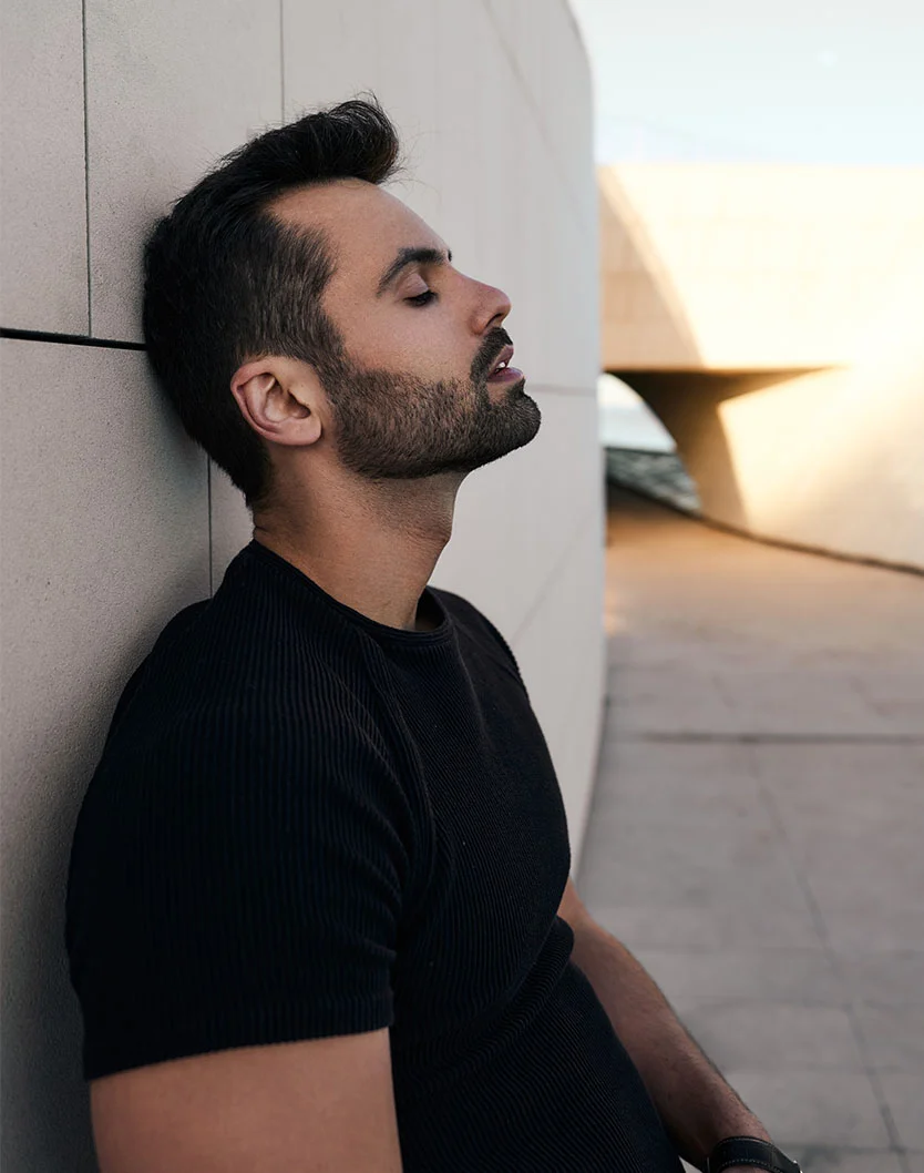 A man with a neatly trimmed beard and dark hair wearing a black ribbed shirt leans against a light-colored, angled wall with his eyes closed, chin slightly lifted. - Male Submental Liposuction Chin Lipo in Orlando Park, IL