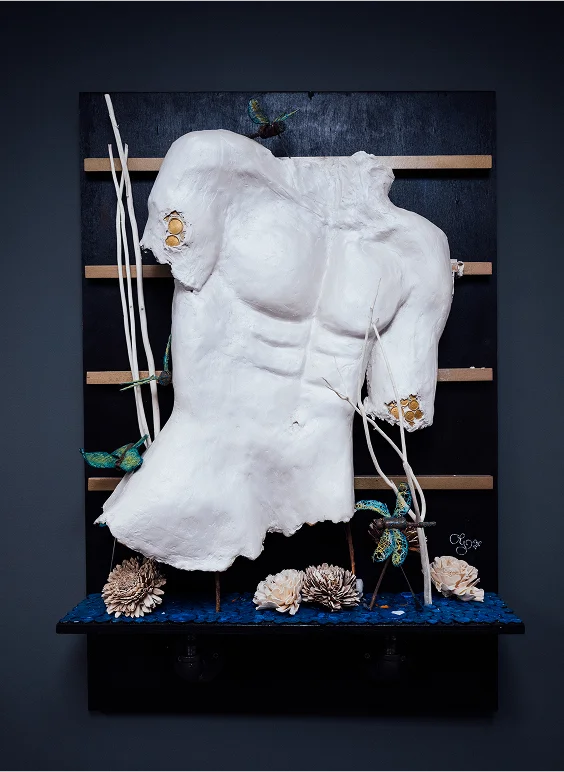 A mixed-media art piece featuring a broken white male torso sculpture mounted on a black and wood backing. The piece includes decorative elements like small artificial hummingbirds, white branches, white flowers, and blue accents at the base.