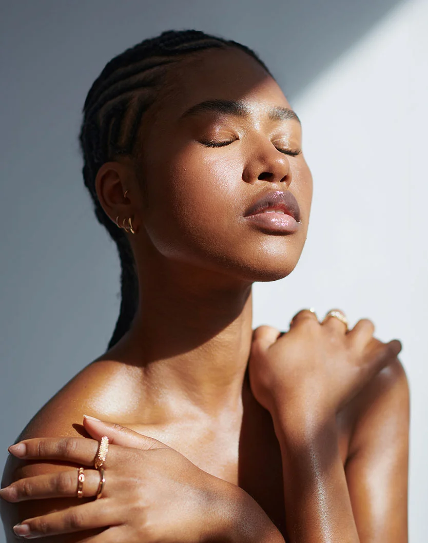 A young woman with glowing, dark skin and intricate cornrows stands in direct sunlight with her eyes closed and arms crossed over her bare shoulders. She is wearing several gold rings. - Microneedling in Orlando Park, IL