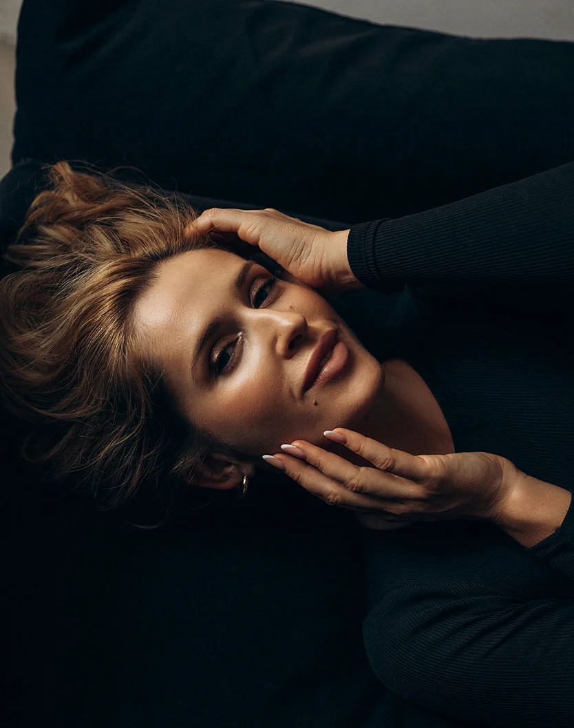 A portrait of a woman with blonde hair lying on a dark surface, wearing a black top, framing her face with both hands while looking at the camera. - Mole Cyst Removal in Orlando Park, IL