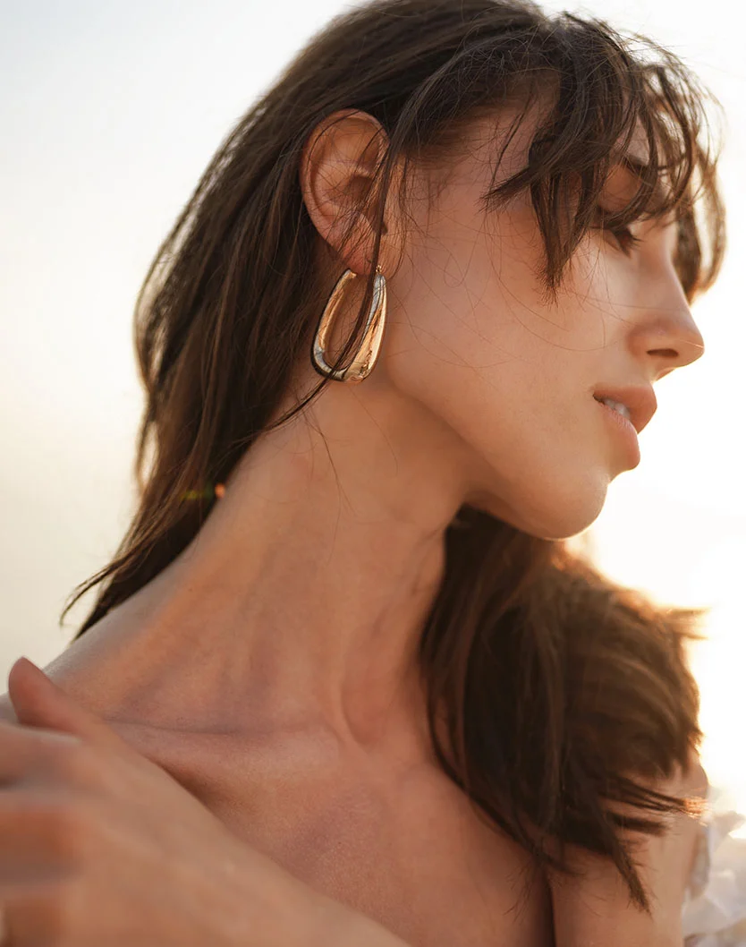 A close-up side profile of a woman with long dark hair and bangs, wearing a large gold hoop earring, illuminated by soft golden light. - Necklift in Orlando Park, IL