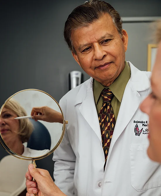 Rajendra Shah, MD, a man wearing a white lab coat, green shirt, and patterned tie, is looking down at a female patient holding a hand mirror. The patient's face is reflected in the mirror, where another hand is gently touching her face.