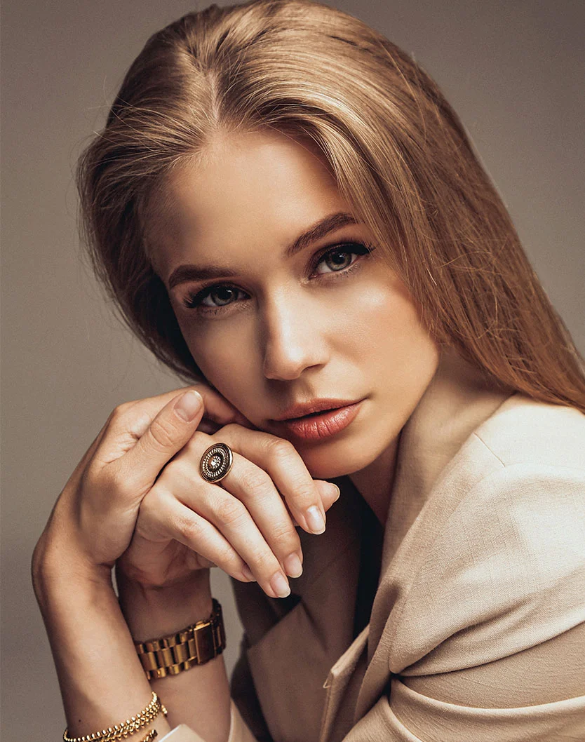A young woman with long, blonde hair, wearing a beige blazer. She is seated, looking directly at the camera with her hands clasped together near her chin. She is wearing a gold watch, two gold bracelets, and a ring with a large black and gold design. The background is a solid, light gray. - Rhinoplasty in Orlando Park, IL