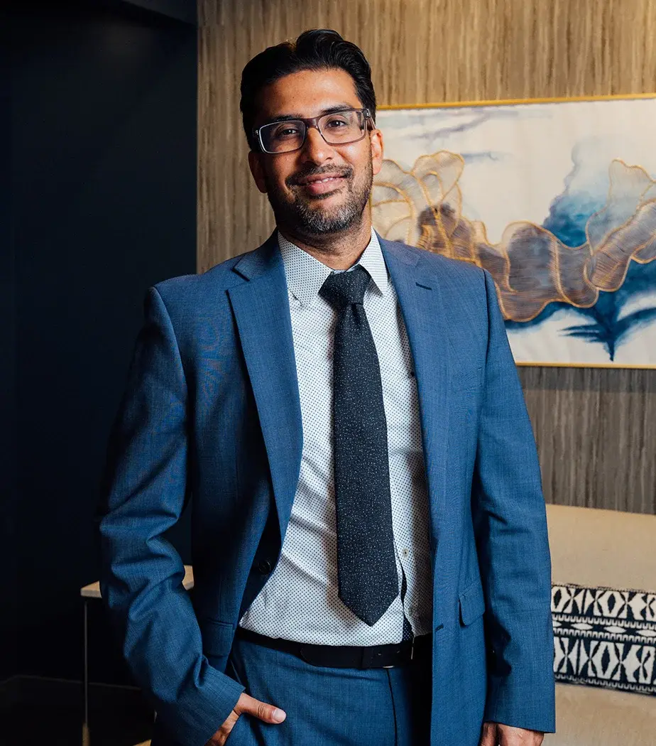 A portrait of Samir Shah, MD, a smiling man with dark hair, glasses, and a beard, wearing a blue suit, patterned white shirt, and dark tie. He is standing indoors in front of a textured wall with abstract art.
