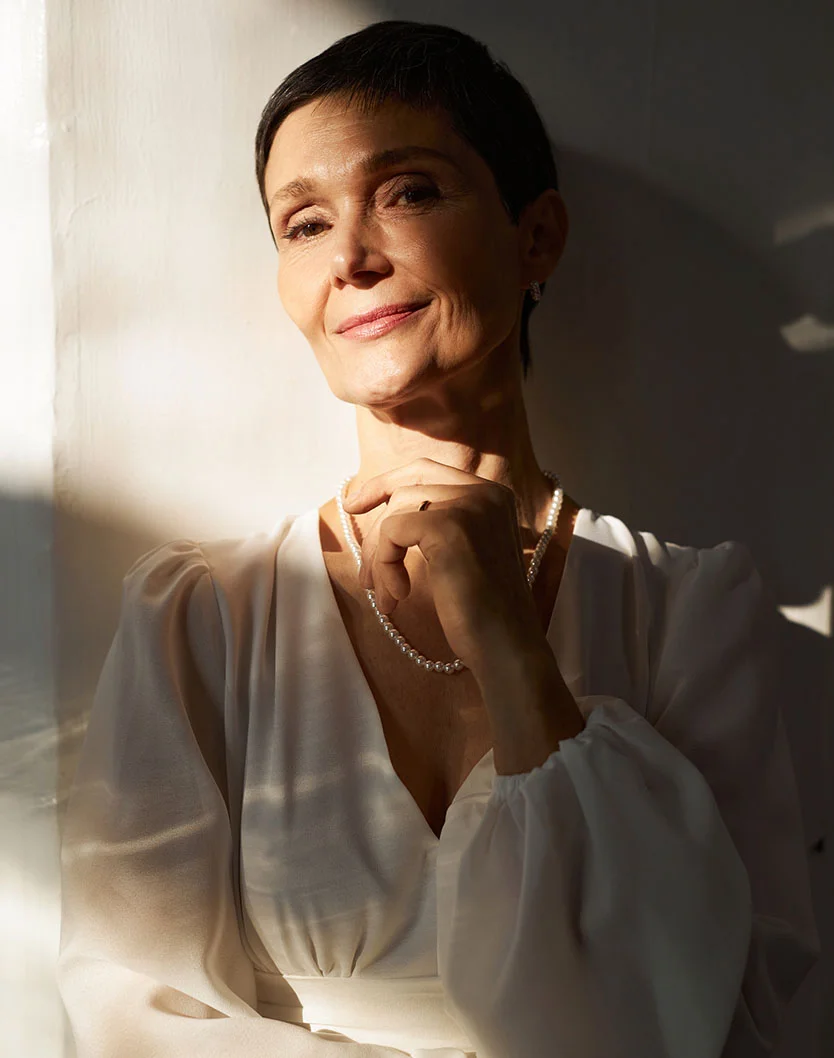 A confident, middle-aged woman with short dark hair, wearing a white v-neck top and a pearl necklace, stands in dramatic sunlight with her hand touching her chin. - Skin Cancer in Orlando Park, IL