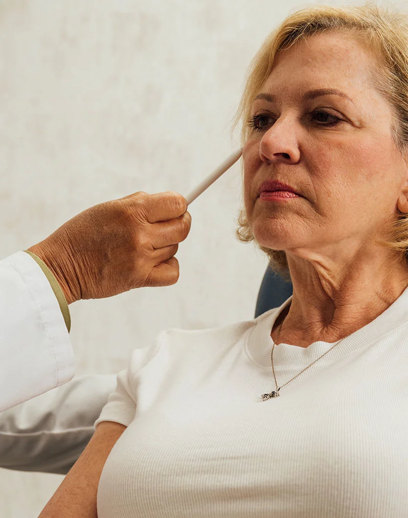 A mature woman with blonde hair sits in a chair while a doctor, wearing a white coat, uses a medical tool to examine the skin near her eye. - Skin Lesions in Orlando Park, IL