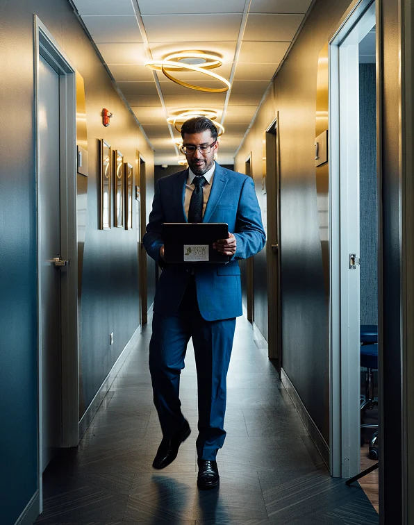 Samir Shah, MD, a man wearing a blue suit, glasses, and a dark tie, is walking down a hallway while looking at a tablet he is holding. The hallway has gray walls, wood-patterned flooring, and distinctive curved gold lighting fixtures.