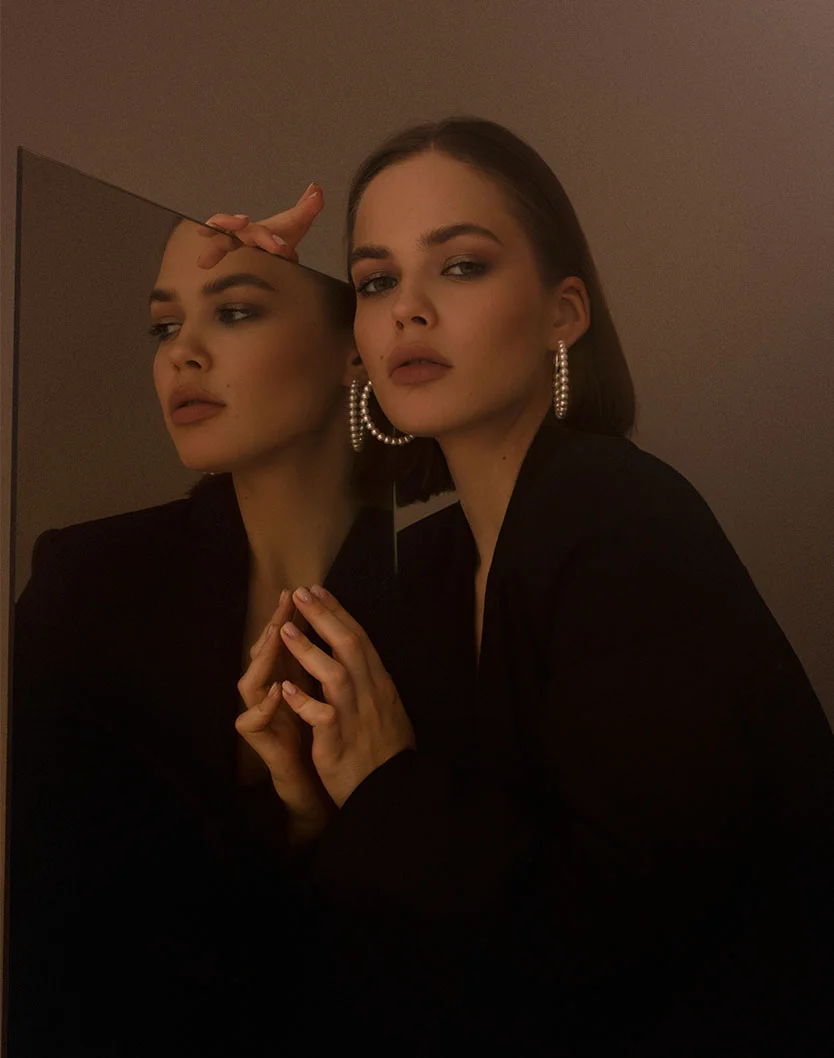 A striking portrait of a young woman with short dark hair and pearl hoop earrings, wearing a black blazer, looking into a mirror and touching her reflection. - Submental Liposuction Chin Lipo in Orlando Park, IL