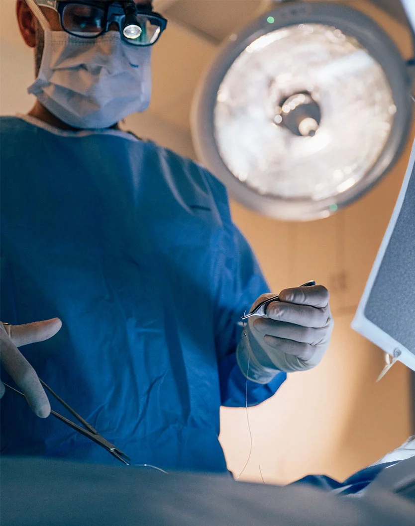 A close-up of Samir Shah, MD, wearing a surgical mask, glasses, and a blue surgical gown, holding a needle and thread with surgical tools while performing a procedure under a bright overhead light. - Tummy Tuck in Orlando Park, IL