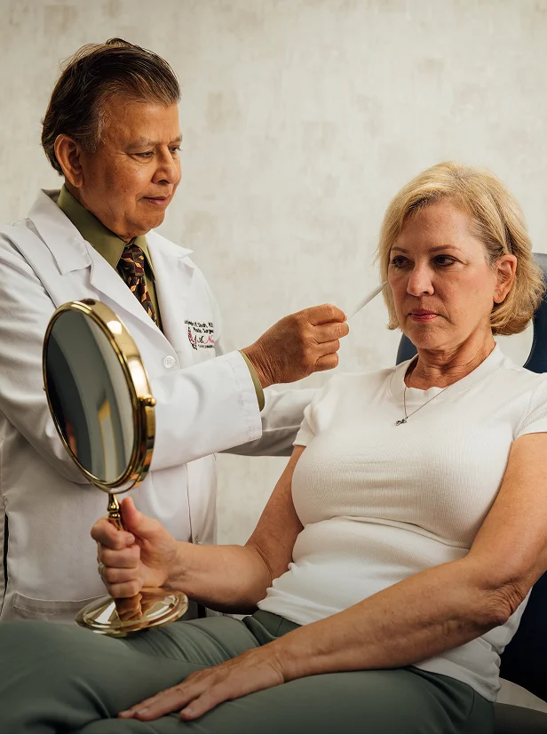Rajendra Shah, MD, a man wearing a white lab coat, green shirt, and tie, is standing beside a female patient seated in a chair. He is holding a white instrument near her temple while she looks at her face in a small, handheld gold mirror.